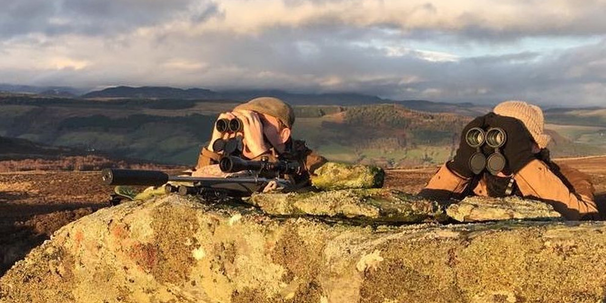 2 deer stalkers hiding behind large rock using binoculars